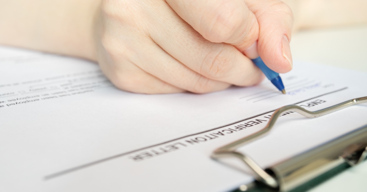 A hand holding a blue pen while writing on an employment verification document attached to a clipboard.