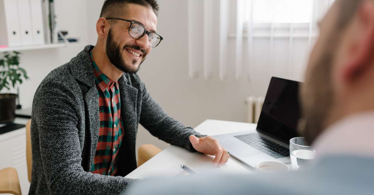 A man with glasses and a sweater sitting behind a desk. His laptop is open and he smiles at the man sitting across from him.