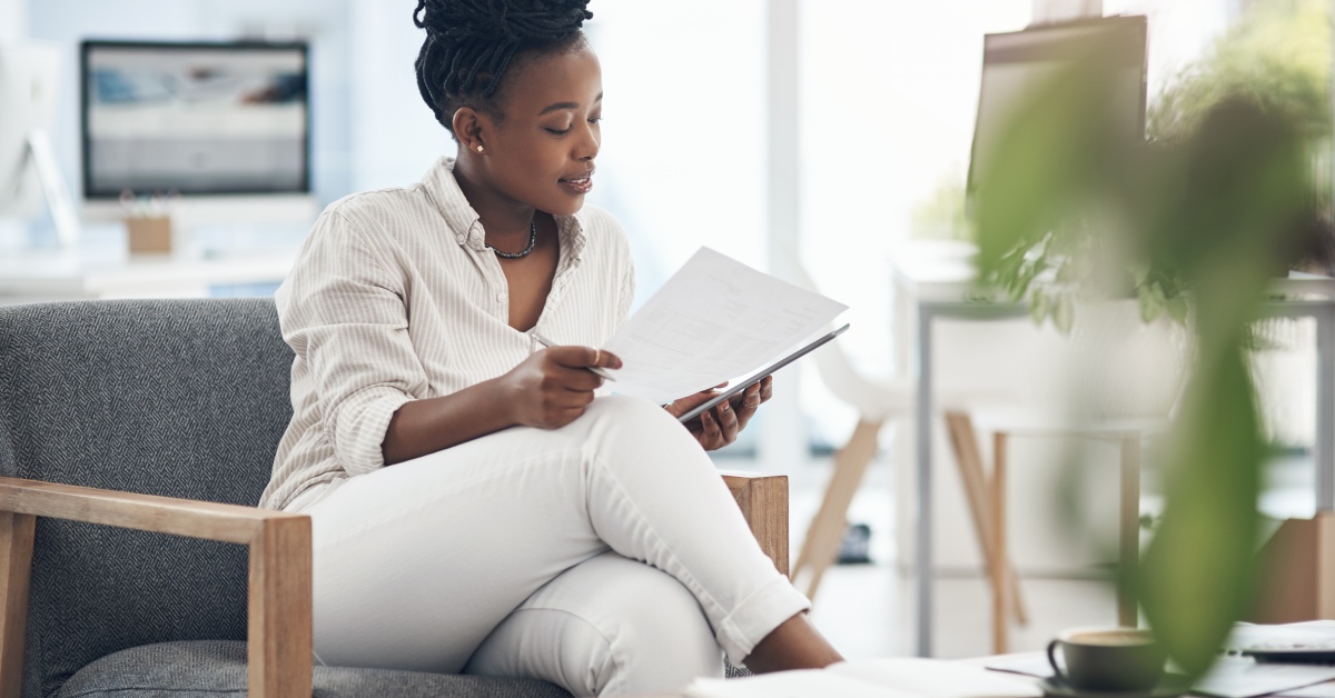 A woman in white clothing is seated and intently reviewing a set of documents while holding a pen in her hand.