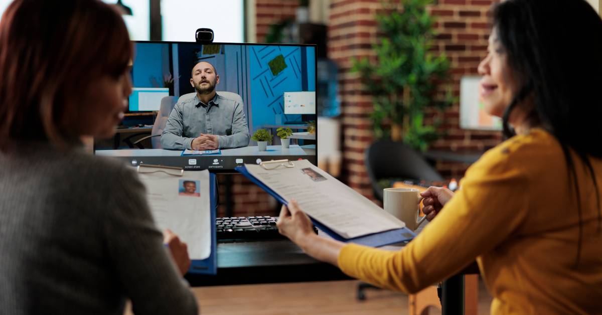 Two women conduct a virtual meeting with a man. The women are holding papers describing job applicants.
