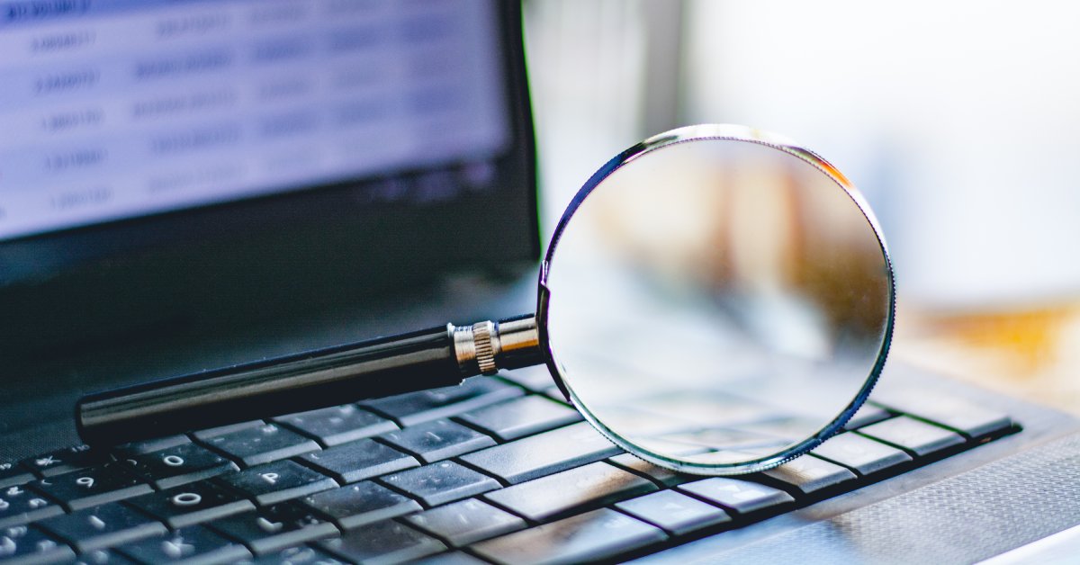 A large magnifying glass lies on its side on top of a black laptop keyboard. The laptop's screen is blurred.