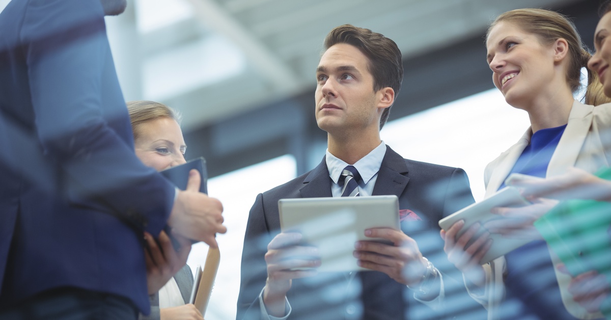 A close-up showing a man wearing a navy blue business suit standing in a group of people while holding a tablet.
