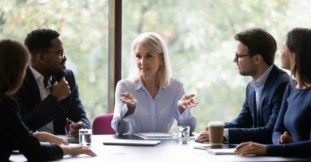 An older woman wearing a blue shirt sits at the head of a table while talking to people in business suits.