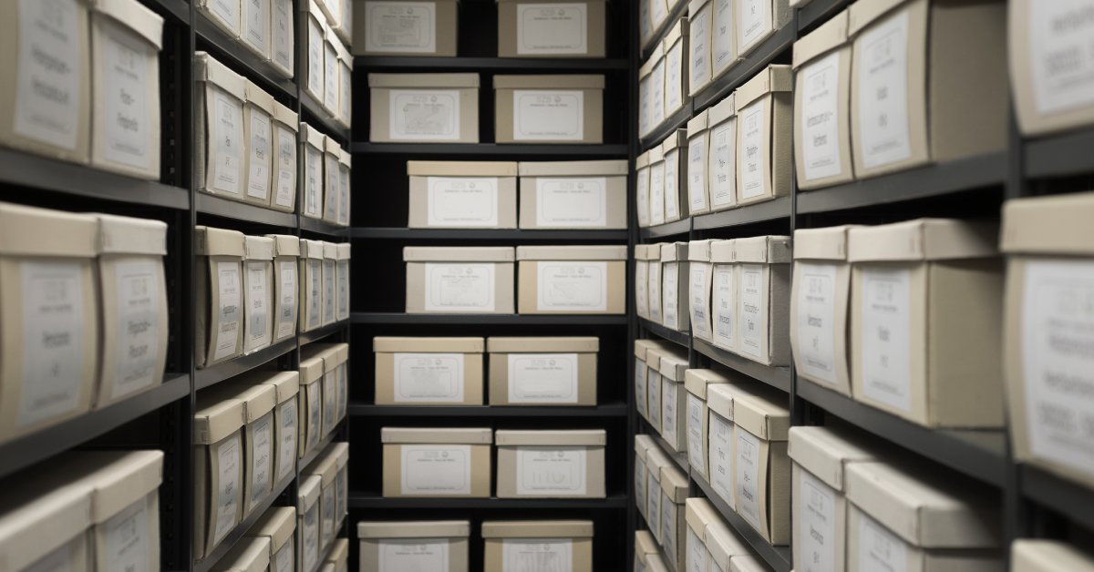 A narrow room lined with dark colored shelves containing stacks of light tan boxes with white labels.