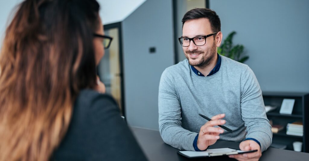 A man wearing glasses and a grey sweater is holding a clipboard and smiling at a woman sitting across the desk.