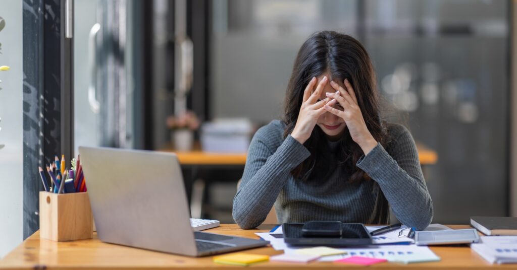 A young woman wearing a grey sweater is sitting at a desk in front of a laptop with her head in her hands.