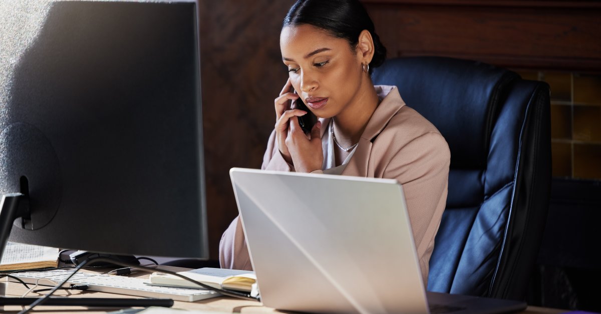 A serious-looking woman wearing a pink jacket sits at a desk in front of a large computer while talking on the phone.