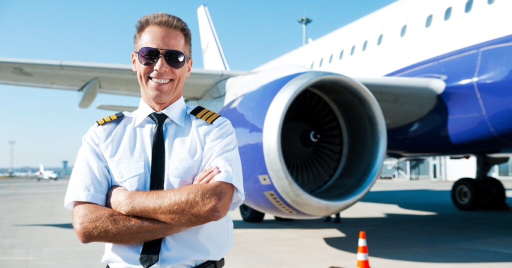 An older man wearing aviator sunglasses and a pilot uniform stands in front of a large blue-and-white airplane.