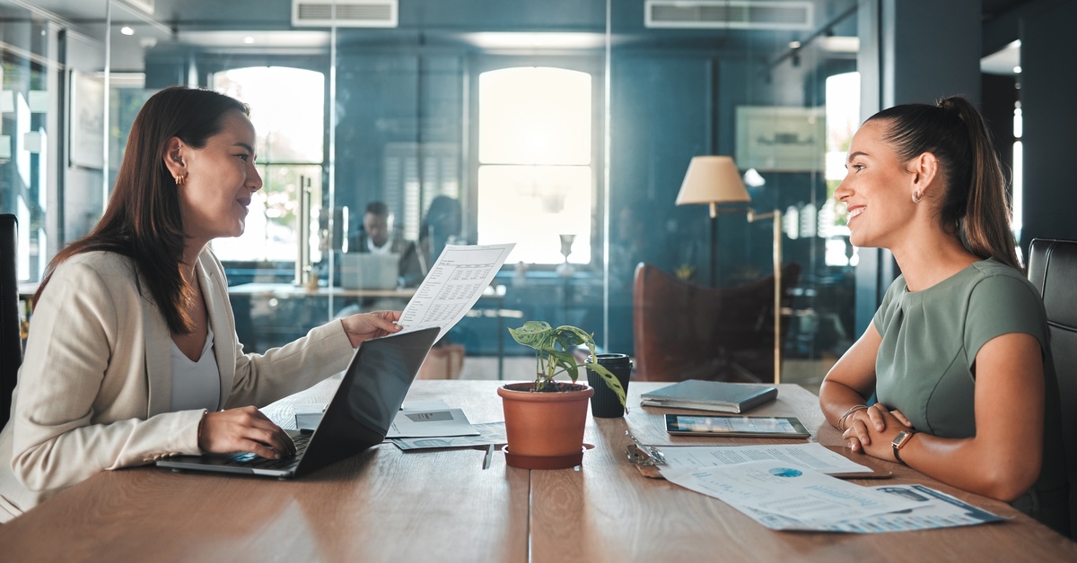 Two women sit across each other at a table in an office. One woman has a laptop and the other has several pieces of paper.