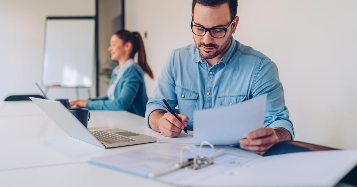 A bearded man wearing glasses is sitting at a white table in front of a laptop and writing on a piece of paper.