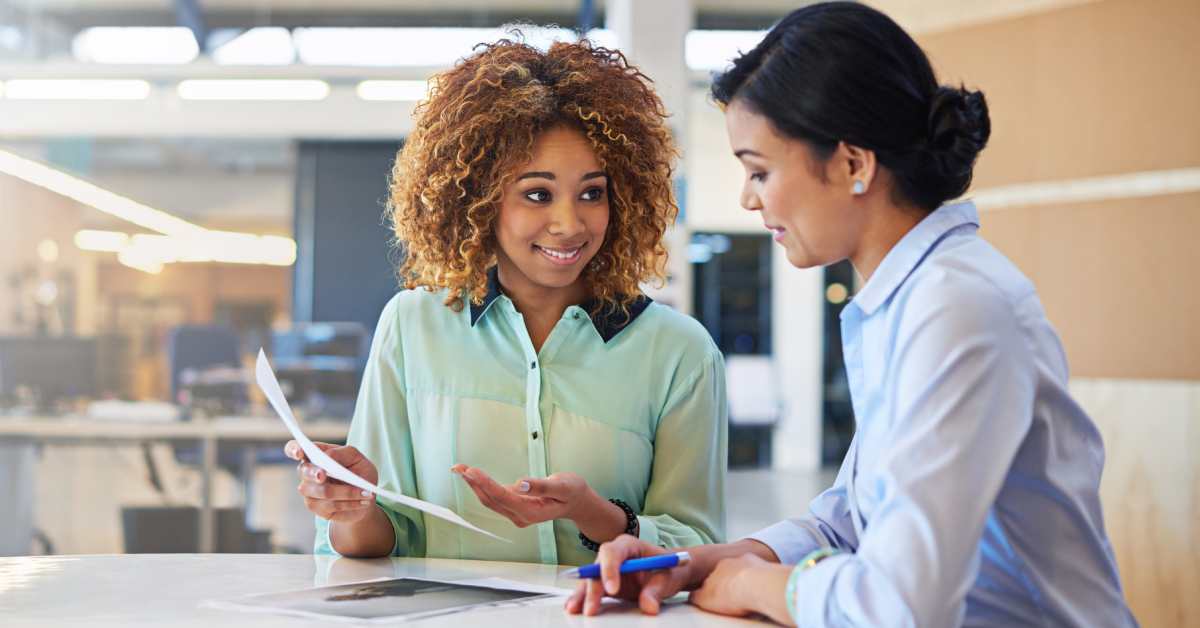 Two women wearing button-down shirts are sitting at a white table in an office and looking at a sheet of paper.