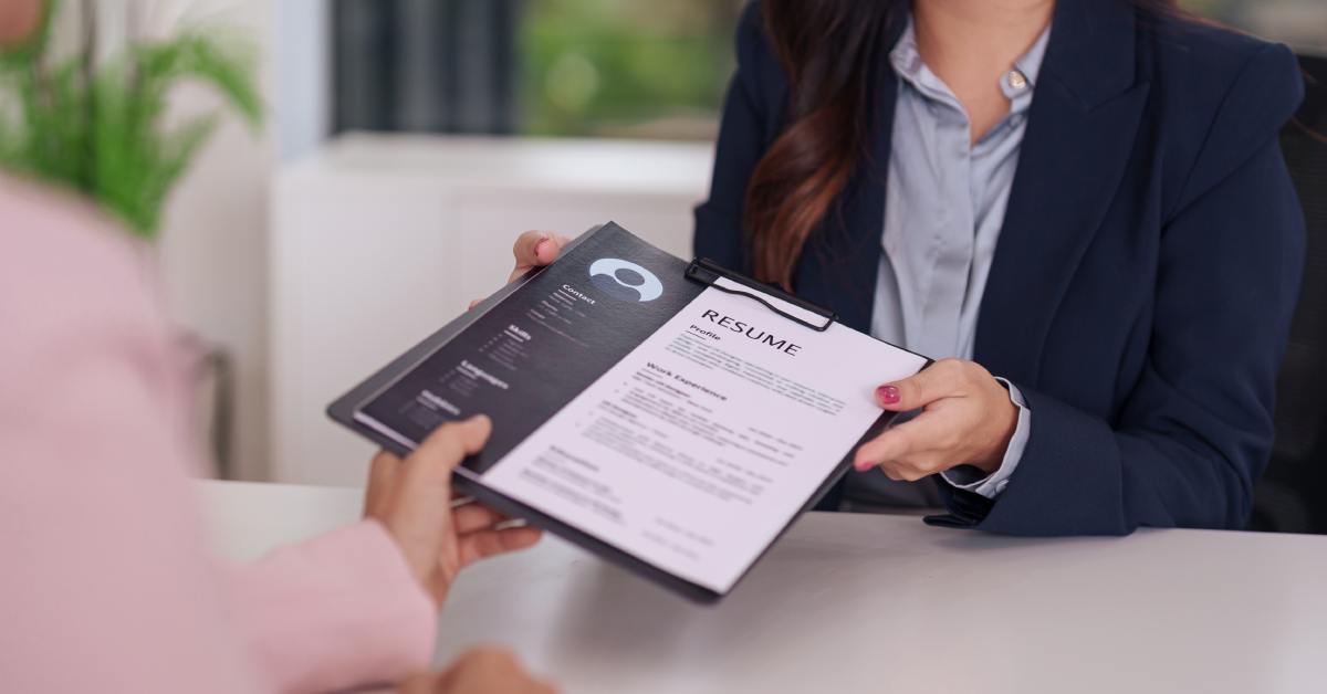 The hands and body of a person as they hand their resume over to a recruiter. The person is seated at a white table.