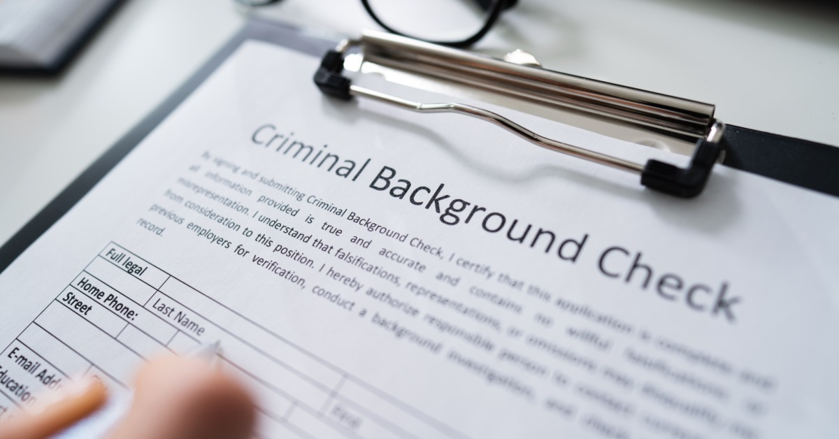 A person filling out a criminal background check form that is attached to a clipboard with a white pen.