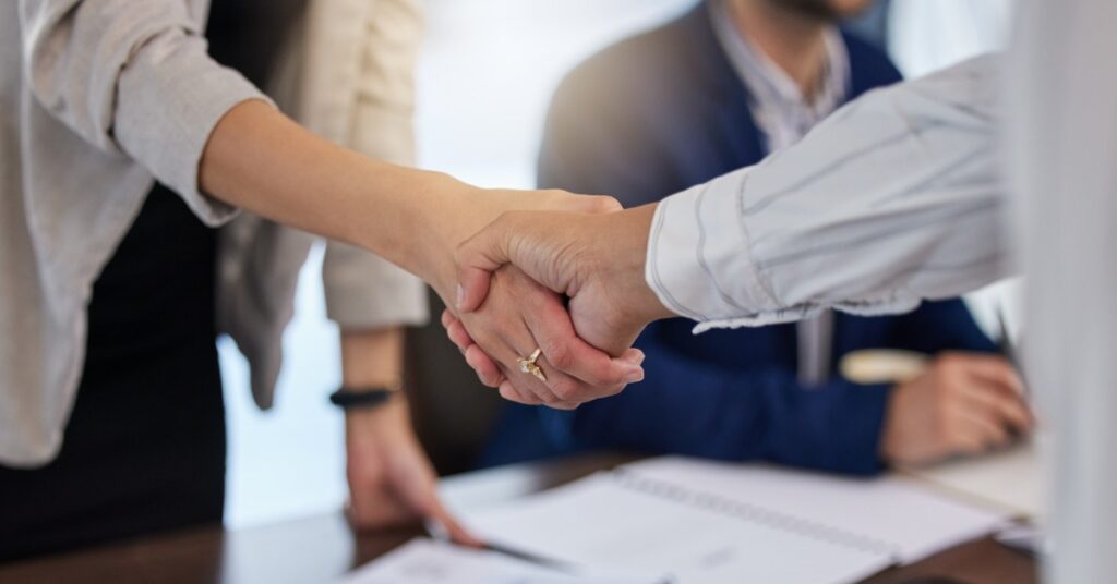 Two individuals in a corporate office shaking hands over a desk. Another person is sitting at the table beside them.