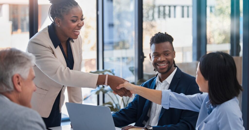 A woman standing and shaking hands with a seated person while two other people sit at a table in an office.