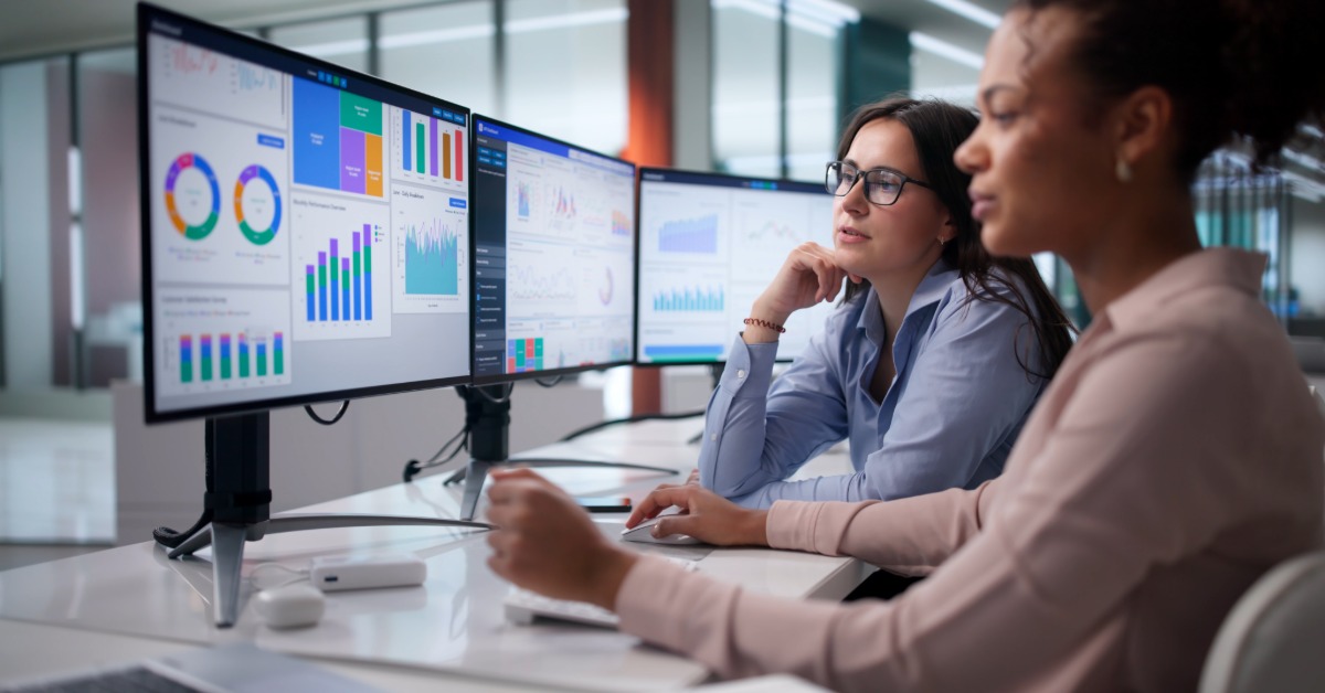 Two women in business-casual attire are sitting at a table, looking at a computer screen displaying charts.