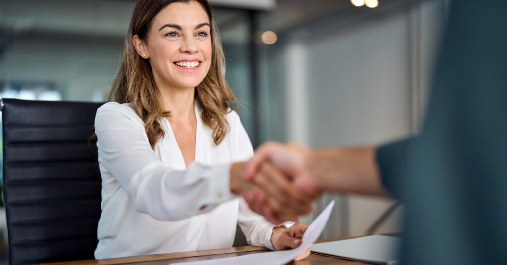 A smiling woman in a white blouse shaking hands with a person across a desk holding papers in an office.