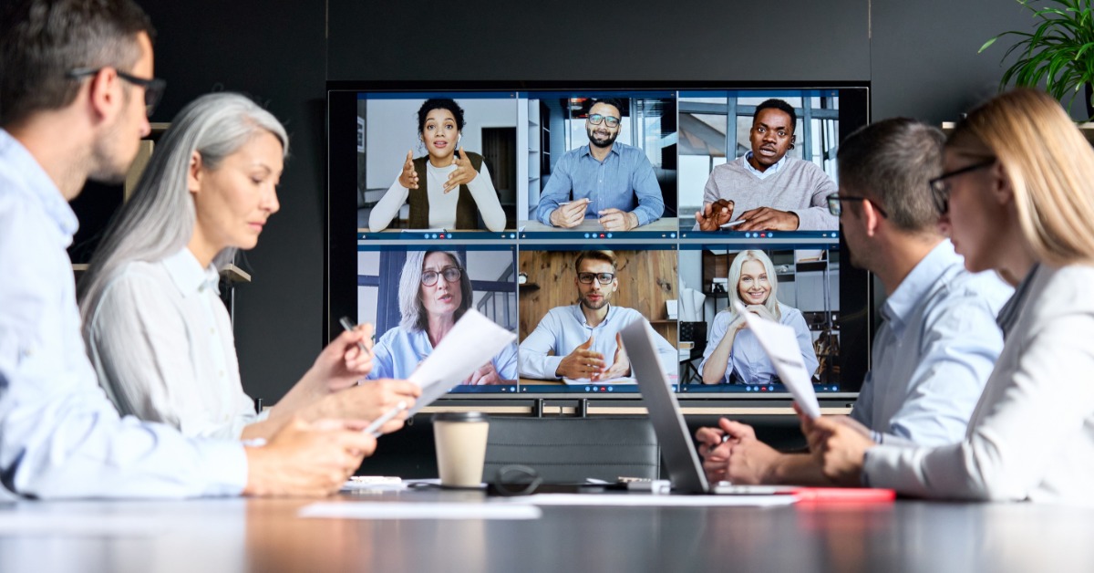 A group of business people sitting around a conference table with other individuals in a virtual meeting on a TV screen.