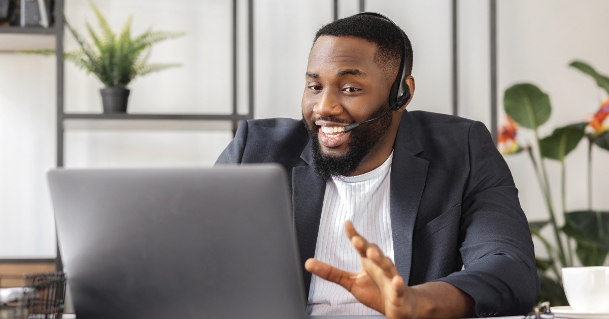 A man working remotely at a desk with his laptop in front of him. He has a headset on for a video call.