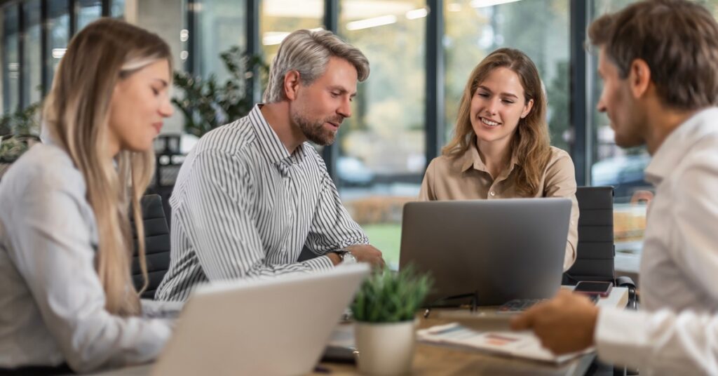Four people sitting around a meeting table with laptops, papers, a small plant, and large windows behind them.