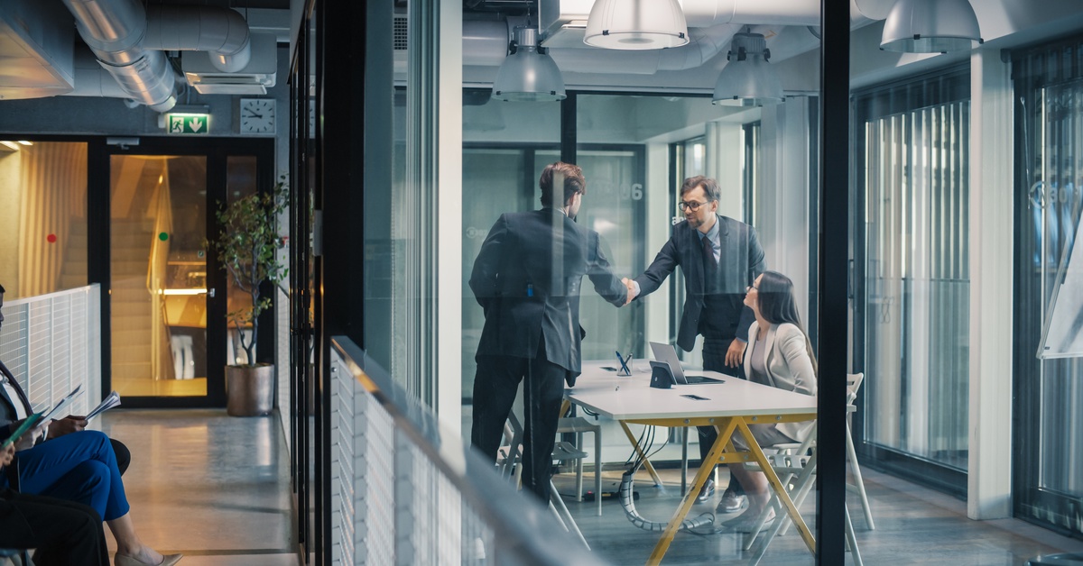 Three people in suits shaking hands in a glass office while another person sits at a table with a laptop.