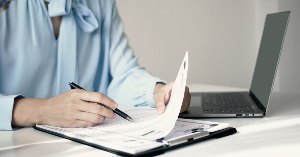 A person in a blue blouse writing on papers clipped to a folder beside an open laptop on a white desk.